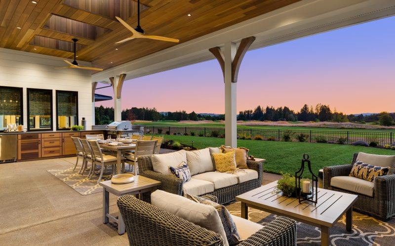 Luxury home exterior at sunset: Outdoor covered patio with kitchen, barbecue, dining table, and seating area, overlooking grass field and trees.